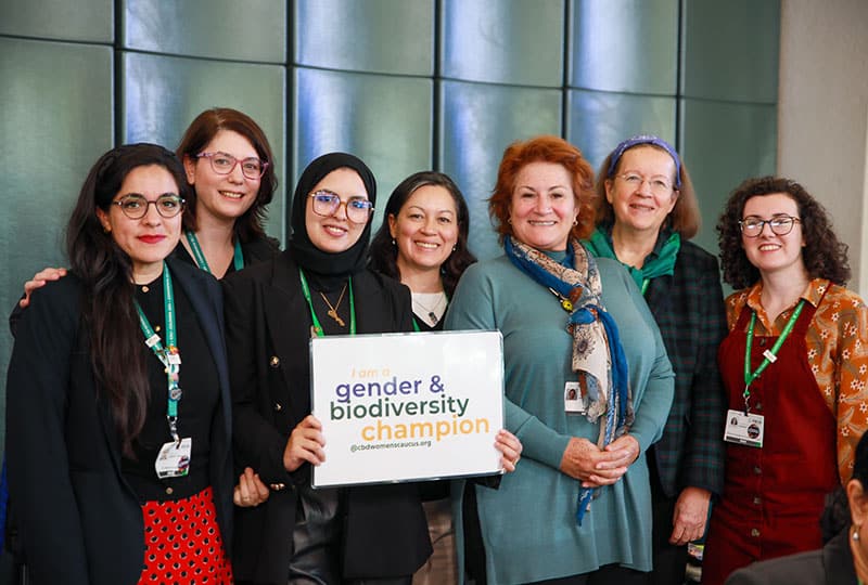 Group photos of members of the CBD Women's Caucus smiling at the camera. One of the women holds up a paper that says